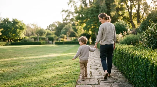 A parent and child walking together along a garden path with a subtle low hedge marking a boundary, symbolizing the line between warmth and limits in parenting